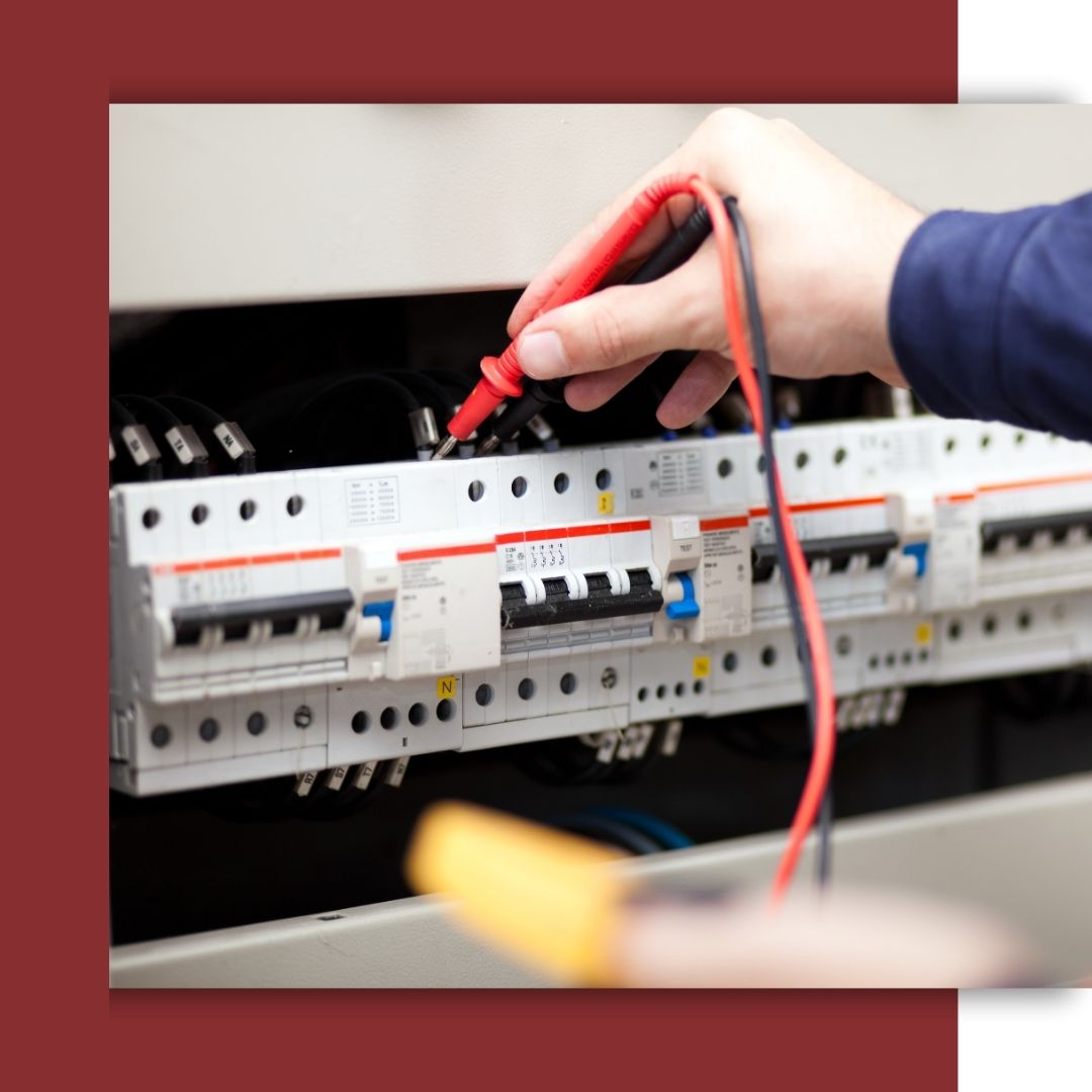 A close-up shot of an electrician's hand using a multimeter to test a commercial circuit breaker panel.
