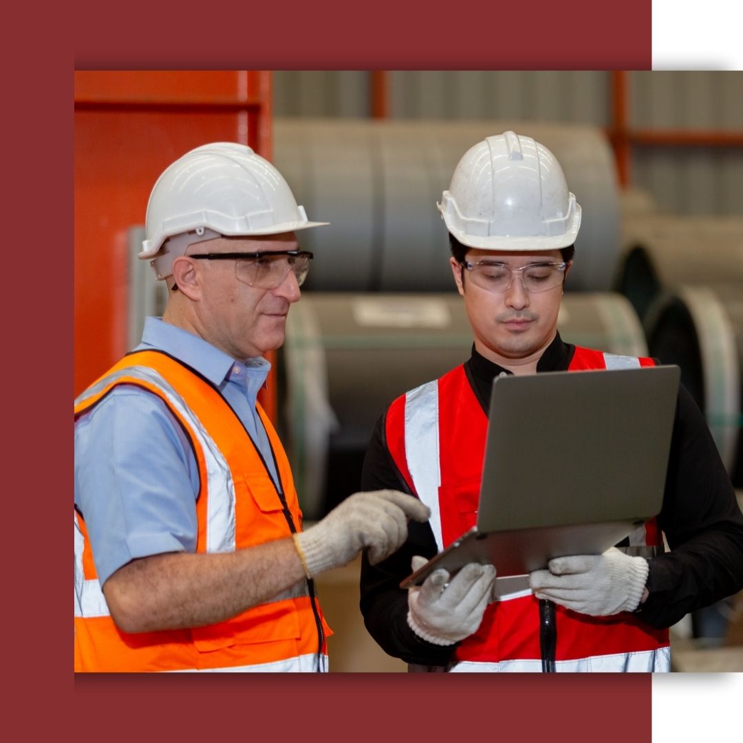 Two commercial electricians in safety vests, hard hats, and gloves review plans on a laptop in an industrial warehouse setting.