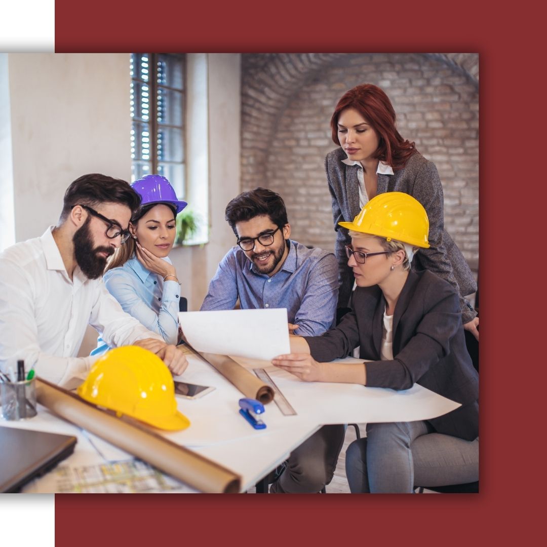 A group of engineers and project managers, some wearing hard hats, gather around an office table to review architectural or electrical blueprints.