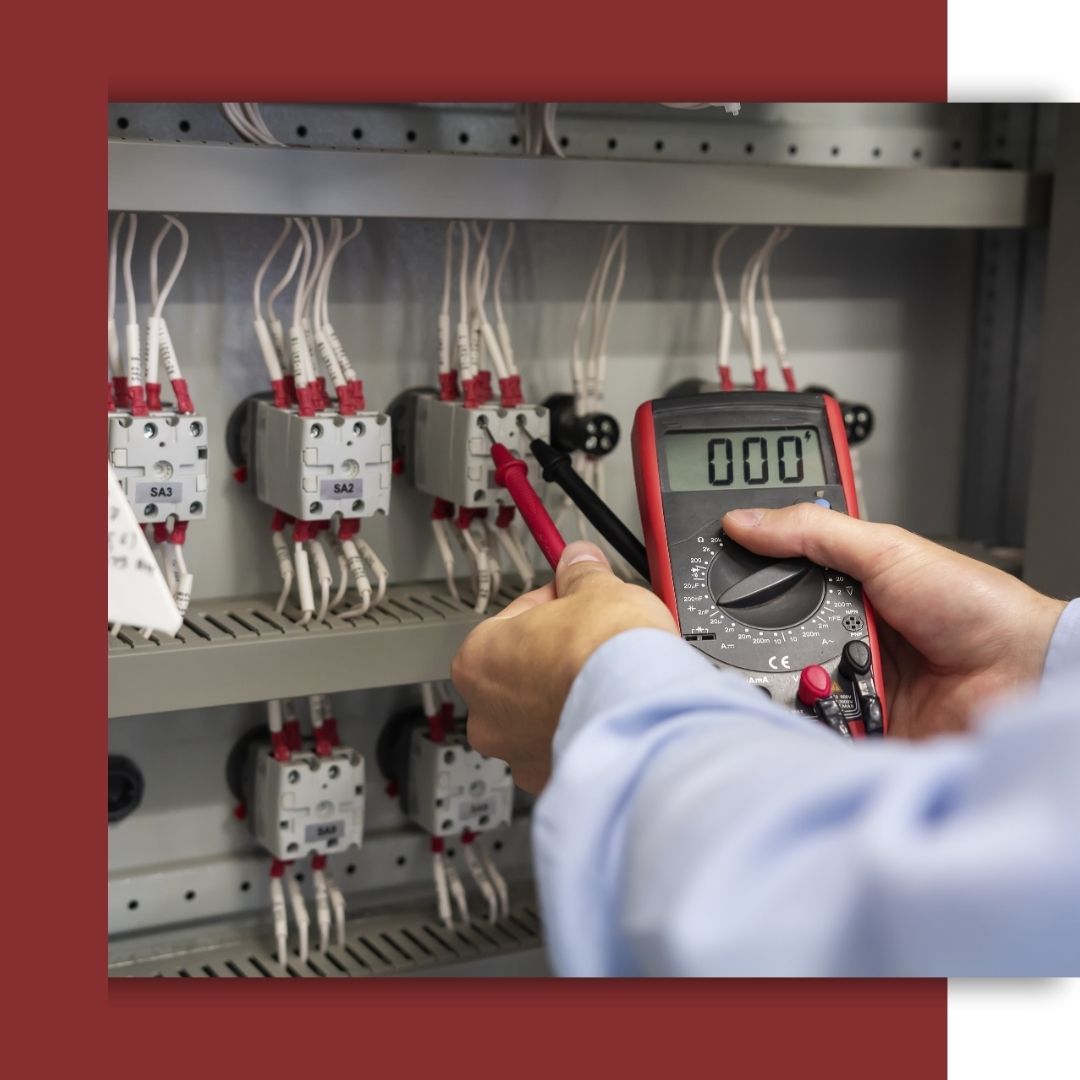 A close-up of an electrician's hands using multimeter probes to test the wiring and terminals inside a complex industrial control panel.