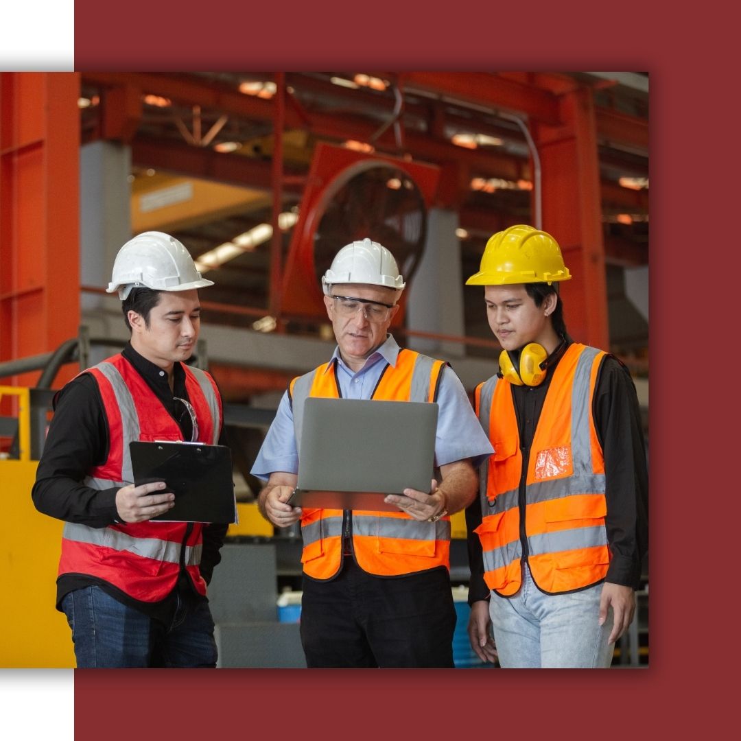 A senior electrical engineer in a hard hat and safety vest uses a laptop to instruct two younger technicians at a large industrial or manufacturing facility.