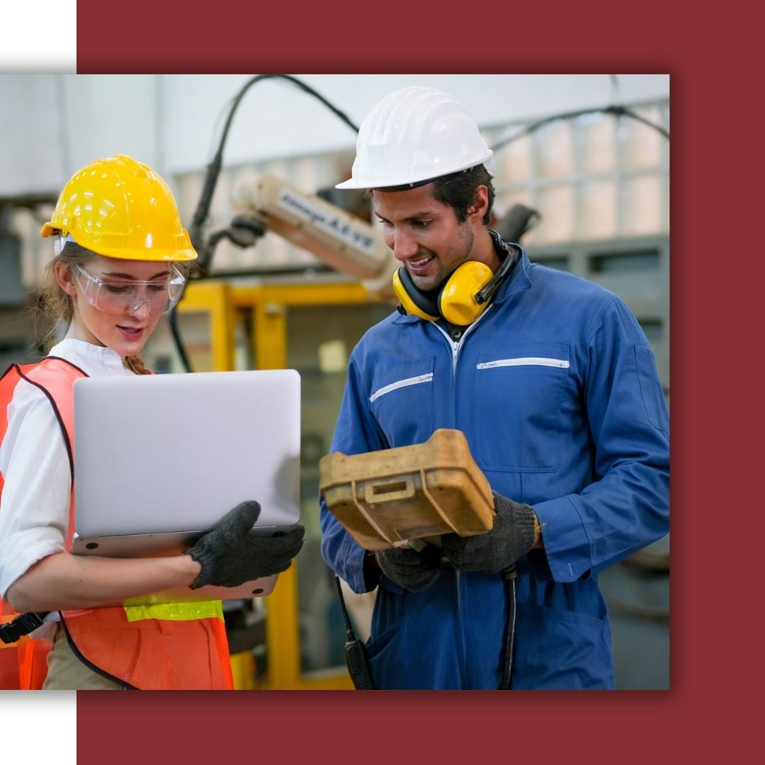 Two industrial technicians, a man and a woman, wearing hard hats and safety gear, smile while collaborating with a laptop and a large handheld control unit in a factory.